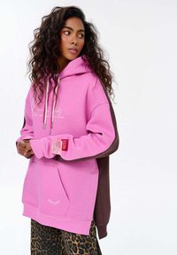 Růžová mikina s hnědými akcenty, se přední kapsou, šňůrkami a ozdobným šitím. Nositá přes sukni s leopardím potiskem.