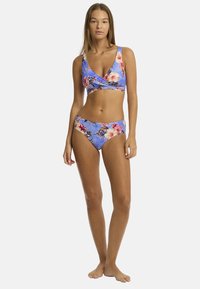 Model staat in een bloemenbikini met een blauwe achtergrond. Ze heeft lang haar en staat tegen een witte achtergrond.
