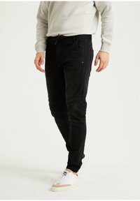 Pantalones negros entallados de mezclilla, con bolsillos laterales, detalles de costura y cintura con cordón, combinados con zapatillas blancas.