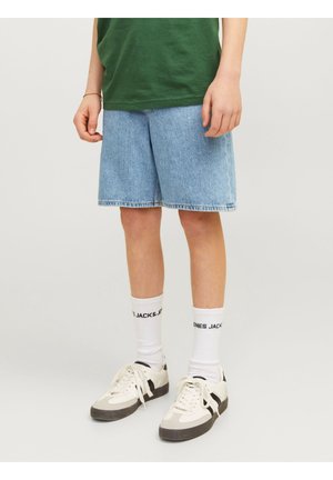 Person iført lyseblå denimshorts, hvide sokker med sort tekst, hvide sneakers med sorte accenter og en grøn skjorte.