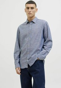 Cămașă lungă de culoare albastru deschis, confecționată din țesătură împletită, cu guler cu nasturi și nasturi pe față, asortată cu blugi din denim închis.