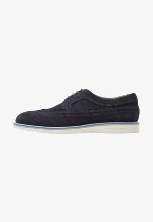 Zapato brogue de piel de ante azul marino para hombre con detalles perforados y suela de goma blanca con ribete azul.
