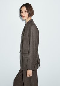 Blazer marrone su misura con un design aderente e due bottoni. Presenta un colletto a rever e una tasca sul petto. Texture liscia.