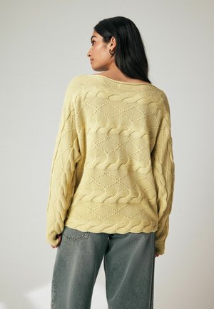 Geel gebreide trui met een kabelpatroon, met een losse pasvorm en brede halslijn. Gepaard met grijze high-waisted jeans. Textuurafwerking.