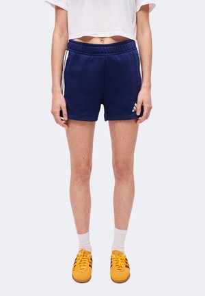 Person iført marineblå træningsshorts med hvide striber, en crop-top hvid T-shirt, hvide crew-sokker og gule sneakers, der står mod en grå baggrund.