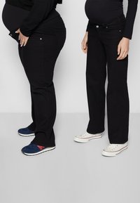 Dois pares de jeans pretos em figuras grávidas, um com um design justo e o outro com um corte reto. Ambos são usados com tênis.