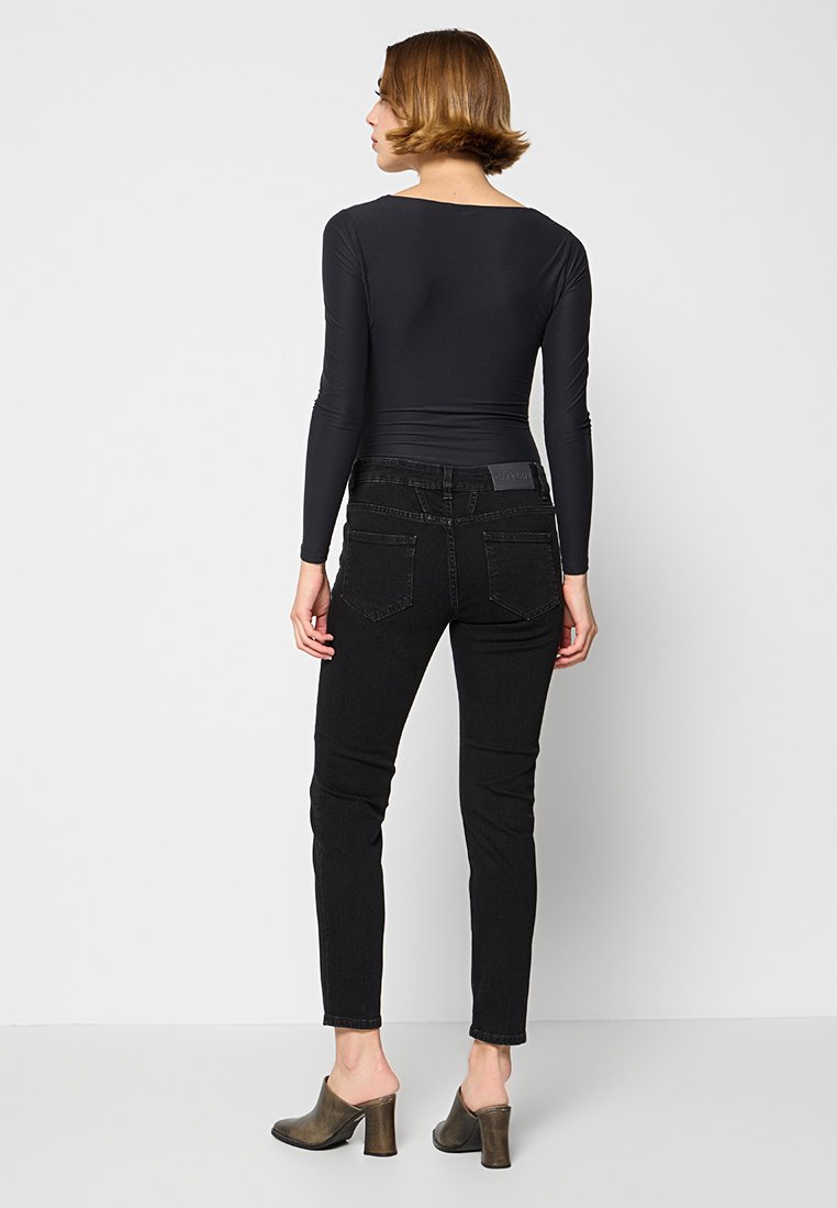 Frau mit kurzen braunen Haaren, die ein enges schwarzes Langarmoberteil, schwarze hochtaillierte Jeans und braune High-Heels trägt und mit dem Rücken steht.