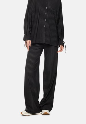 Person wearing loose black pinstripe pants and matching button-up shirt with white sneakers, standing against a light background.