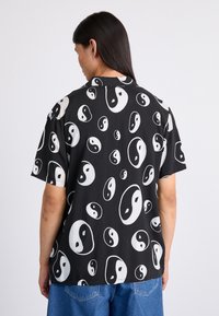 Chemise à manches courtes en tissu noir avec un motif yin-yang blanc répétitif. Coupe décontractée et design col avec un ourlet droit.
