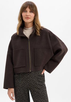 Braune, cropped Jacke mit hohem Kragen, Frontreißverschluss und zwei großen Taschen. Kombiniert mit gemusterten Stoffhosen. Glatte Stoffoberfläche.