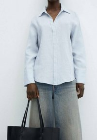 Chemise en lin bleu clair avec un col, patte de boutonnage, et manches longues. Associée à un jean large bleu clair et un sac fourre-tout noir.