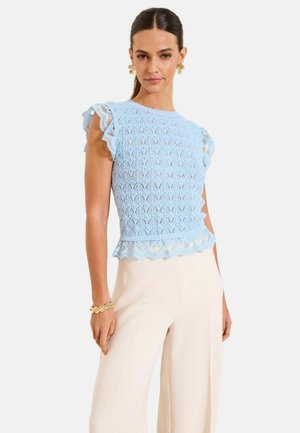 Frau trägt ein hellblaues ärmelloses Spitzentop mit Rüschen, cremefarbene Hosen mit weitem Bein, goldenes Armband und goldene Ohrringe, steht mit der Hand in der Hüfte.