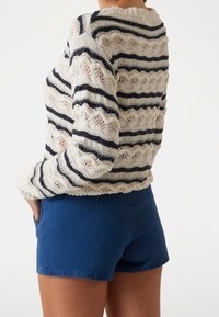 Pletený svetr v krémové barvě s námořnickými pruhy, zdobený zvlněným vzorem. Kombinován s modrými šortkami, které mají volný střih a kapsy.