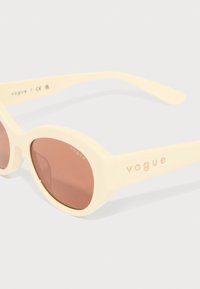 Slnečné okuliare v krémovej farbe s oválnymi šošovkami a lesklým povrchom. Na ramienku je umiestnené branding 'vogue' v ružovom zlate. Rám s textúrovanými akcentmi.