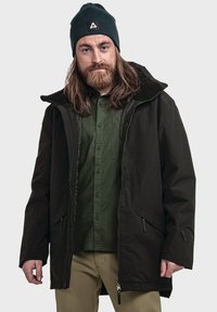 Schwarze wetterfeste Jacke mit hohem Kragen, Frontreißverschlüssen und zwei seitlichen Taschen, über einem grünen Hemd mit Knopfleiste getragen. Dunkle Strickmütze auf dem Kopf.