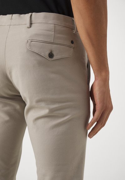 Beige Baumwollhosen mit schlanker Passform, einer Gesäßtasche mit Knopf und glatter Textur. Die Seitenansicht zeigt detaillierte Nähte.