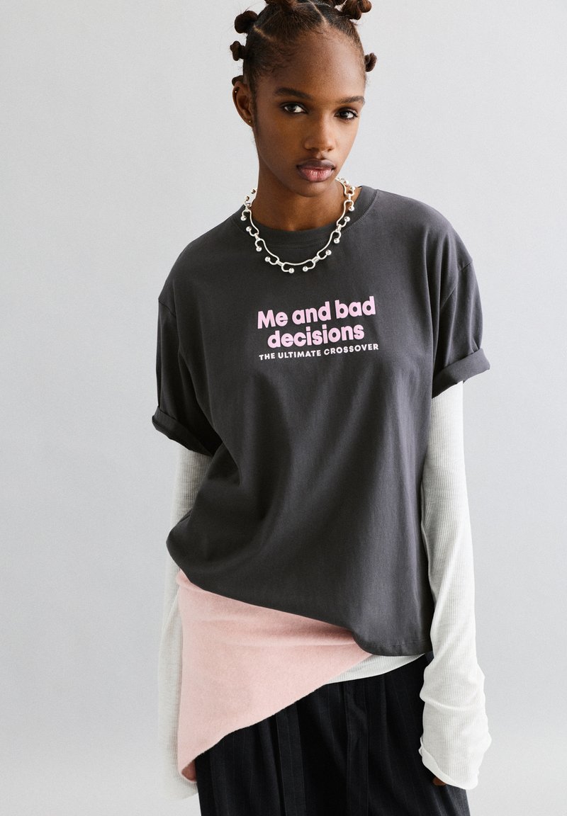 Jonge vrouw met gestyled haar, gekleed in gelaagde kleding en een ketting, met een antracietgrijze t-shirt waarop in roze de tekst "Ik en slechte beslissingen" staat.