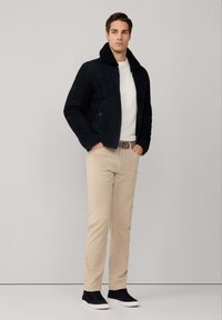 Mann in schwarzer Jacke mit Pelzkragen, weißem Pullover, beigen Hosen und schwarzen Sneakern, der vor einem schlichten grauen Hintergrund steht.