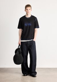 Camiseta preta de manga curta com detalhes azuis apresentando o logótipo "HUGO". Combinada com jeans soltos de ganga escura e uma mala redonda preta.