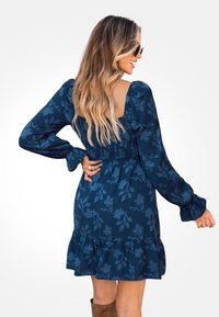 Vestido azul marino con estampado floral, cintura fruncida y mangas largas. Detalle de dobladillo con volantes y tejido texturizado. Usado con botas marrones hasta la rodilla.