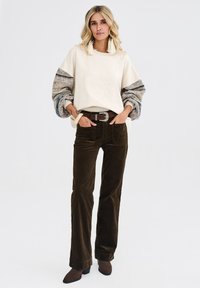 Kremowy sweter z fakturowanymi szaro-brązowymi rękawami, brązowe spodnie z sztruksu i ciemnobrązowe buty na obcasie. Wyróżnia się srebrnym paskiem z klamrą.