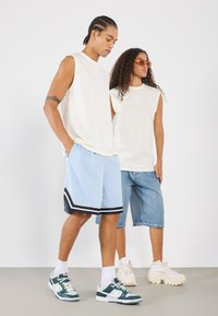 Divi modeļi stāv pretī gaišai fonam. Vīriešu modelis valkā baltu bezprofila kreklu un gaiši zilo sporta šortu komplektu ar melniem akcentiem. Sieviešu modelis valkā baltu bezprofila topu un džinsu šortus. Abi sporto ikdienišķus apavus.