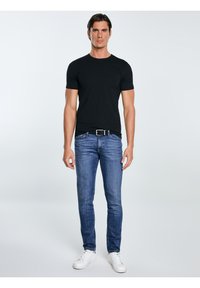 Ein Mann in einem schwarzen T-Shirt, blauen Jeans mit einem schwarzen Gürtel und weißen Turnschuhen steht vor einem schlichten hellen Hintergrund.