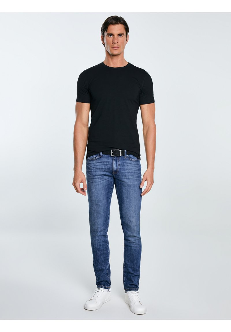 Ein Mann in einem schwarzen T-Shirt, blauen Jeans mit einem schwarzen Gürtel und weißen Turnschuhen steht vor einem schlichten hellen Hintergrund.