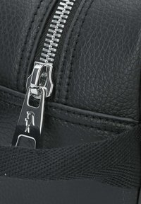 Dettaglio ravvicinato di una borsa in pelle nera texturizzata con una cerniera metallica e un cursore in argento lucido. Include un particolare di un manico intrecciato nero.