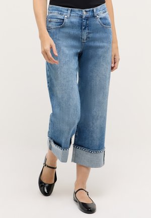 Woman wearing light blue cuffed jeans with pearl detailing paired with black Mary Jane shoes, standing against a plain background.