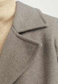 Manteau en laine gris avec col cranté et un petit détail de boutonnière décoratif, superposé à un col roulé beige à côtes. Texture douce visible.