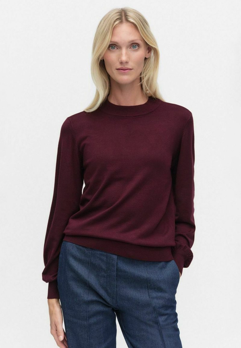 Bordowy sweter z dzianiny z okrągłym dekoltem, długimi rękawami oraz prążkowanymi mankietami i dołem, w zestawieniu z niebieskimi dopasowanymi spodniami.