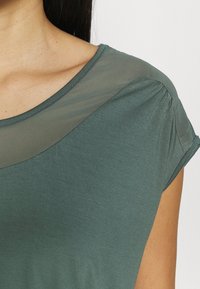 Žena oblečená v zeleném triku s krátkým rukávem, které má průsvitnou síťovanou část přes horní část hrudníku a ramena.