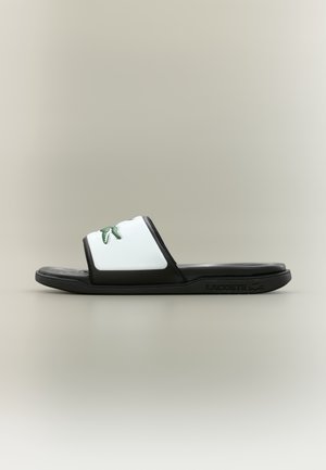 Sandalia tipo chancla Lacoste en blanco y negro con un logo de cocodrilo verde en la correa y suela texturizada sobre un fondo neutro.