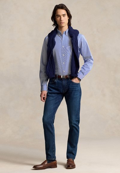 Fiatal férfi kék gombos ingben, sötétkék, kábelmintás pulóvert vállára vetve, kék farmernadrágban, barna övvel és barna mokaszinban, egyik kezét zsebébe téve áll.
