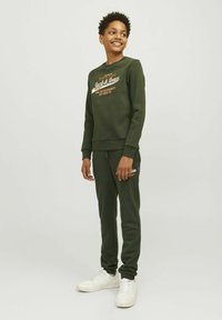 Mørkegrøn sweatshirt og matchende joggers, lavet af blødt stof. Har et logo i kontrasterende farver på brystet og bukserne. Hvide sneakers.