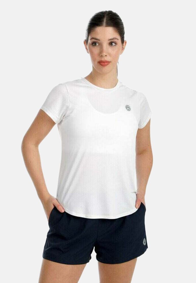 Mujer de cabello oscuro con una camiseta blanca de manga corta y pantalones cortos deportivos negros, de pie con las manos en los bolsillos, mirando ligeramente hacia la derecha.