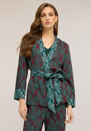 Vrouw draagt een pak met een blauwgroene en kastanjebruine paisley-print, voorzien van een ceintuur, lange mouwen en gouden hangende oorbellen, staand tegen een effen achtergrond.