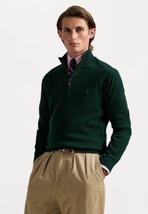Dunkelgrüner Pullover mit Reißverschluss aus weichem Stoff. Getragen über einem hellrosa Hemd mit Kragen und khakifarbenen Hosen. Subtiler Logodetail.