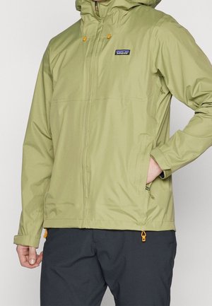 Uomo che indossa una giacca Patagonia verde chiaro con cappuccio e tasche con zip, pantaloni neri, con una mano nella tasca della giacca.