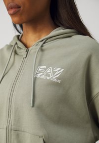 Svijetlozelena zip-up dukserica od meke tkanine s bijelim EA7 Emporio Armani logotipom i vezicama s zaobljenim kapuljačom.