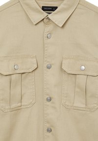Chandail beige à boutons avec deux poches poitrine, boutons-pression argentés et un col classique. Fabriqué à partir d'un tissu durable avec une finition texturée.