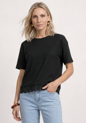 Femme blonde portant un haut noir à manches courtes avec ourlet en dentelle, jean bleu clair, boucles d'oreilles en or et un bracelet marron, posant devant un fond blanc.