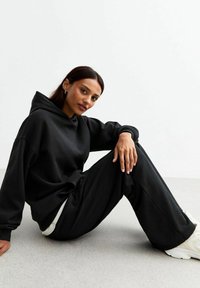 Felpa nera oversize con cappuccio e maniche lunghe, abbinata a pantaloni neri larghi. Entrambi sono realizzati in un morbido tessuto testurizzato.