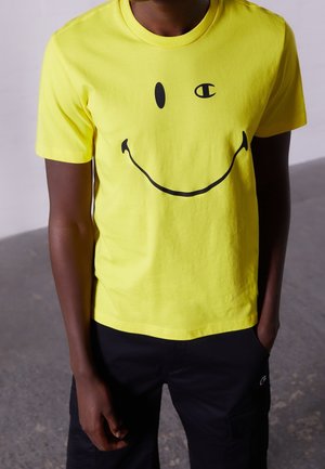 Gul bomuld t-shirt med stort sort smilende ansigt grafik og en lille logo detalje. Kortærmet med rund hals.