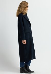 Langer, dunkelblauer Mantel mit einem doppelt knopfartigen Design, strukturierten Revers und zwei Vordertaschen, kombiniert mit enganliegenden Jeans und Stiefeletten.