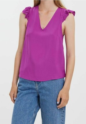 Ærmeløs lilla bluse med flæser på skuldrene og V-hals, båret sammen med blå jeans med høj talje på en stående person.