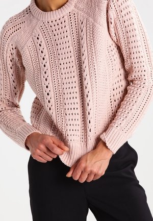 Femme portant un pull tricoté rose pâle avec des trous décoratifs et des poignets côtelés, associé à un pantalon noir, ajustant l'ourlet du pull.