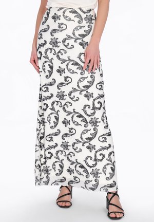Žena v bílé maxi sukni s černým květinovým vzorem a černých vysokých sandálech s pásky, ruce volně podél těla.