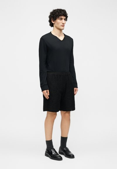 Νέος άνδρας στέκεται φορώντας μαύρο πουκάμισο με μακριά μανίκια, μαύρα δαντελένια σορτς, σκούρες κάλτσες και μαύρα δερμάτινα μοκασίνια, μπροστά σε απλό ανοιχτό φόντο.